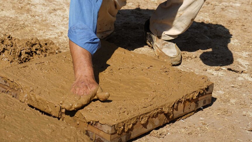 A man runs his hand over a layer of mud packed into a square mould.
