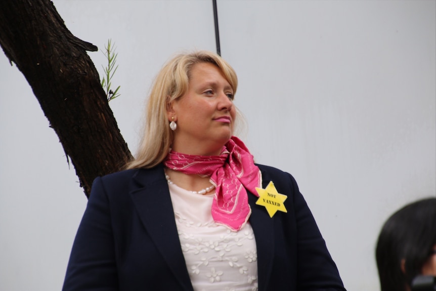 A blonde woman with a pink scarf wears a star of David with the words 'not vaxxed' on her jacket.