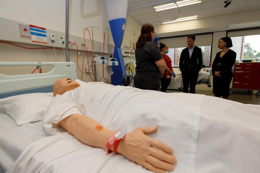 A dummy lies in a bed, a group of people speaking behind it.