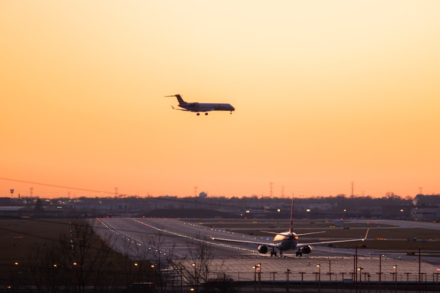A plane approaches an airport runway during a golden sunset. Another waits on the tarmac for take-off.