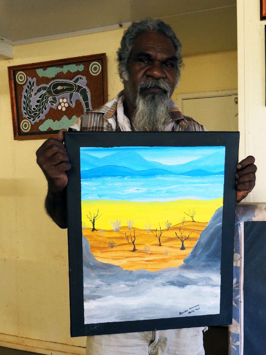 A man photographed holding a set of paintings in the bush, using orange, yellow and blue colors.