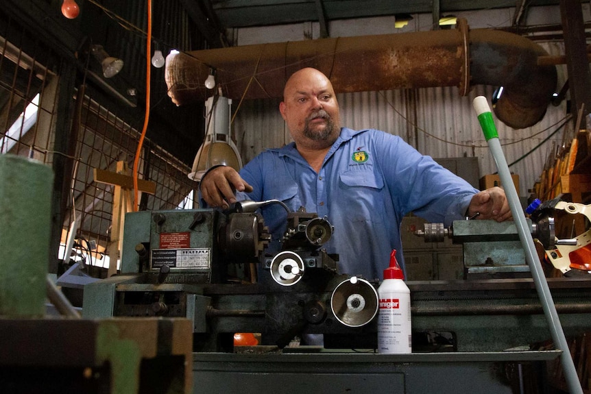 A bald man in a work shirt stands behind metal machinery in a shed full of tools.