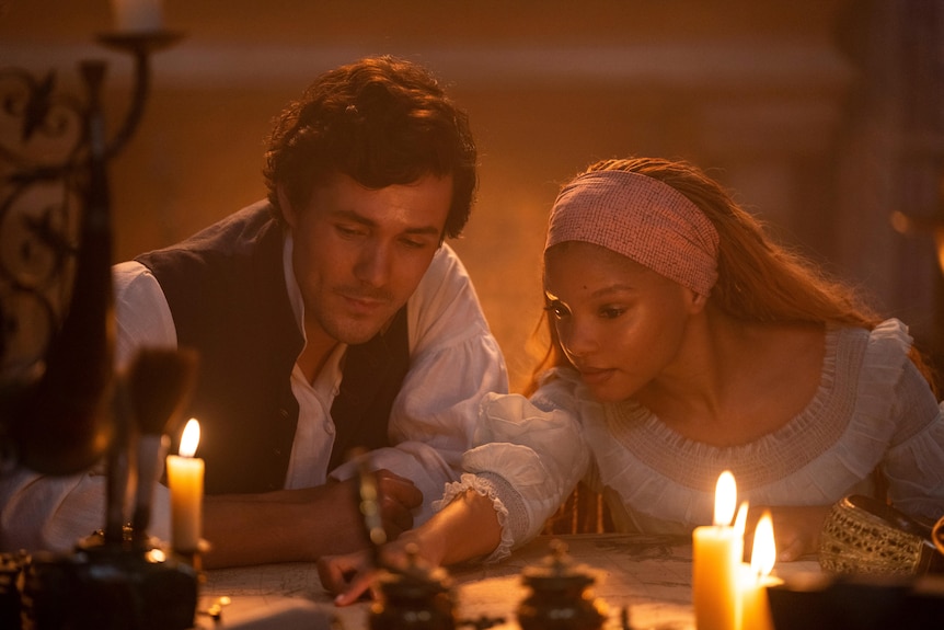 Jonah Hauer-King as Prince Eric and Halle Bailey as Ariel in The Little Mermaid. They're looking at a map in the candlight.