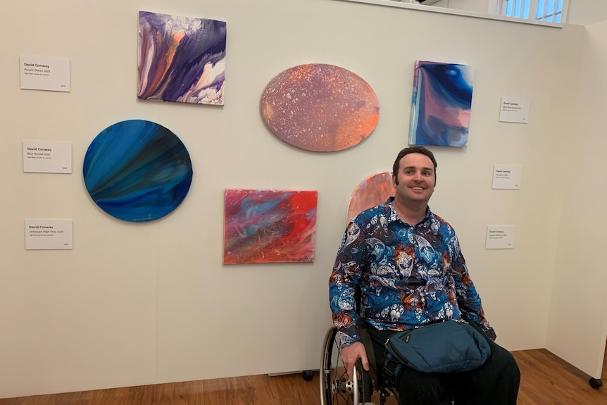 a man smiles at the camera in front of several pieces of colourful abstract art