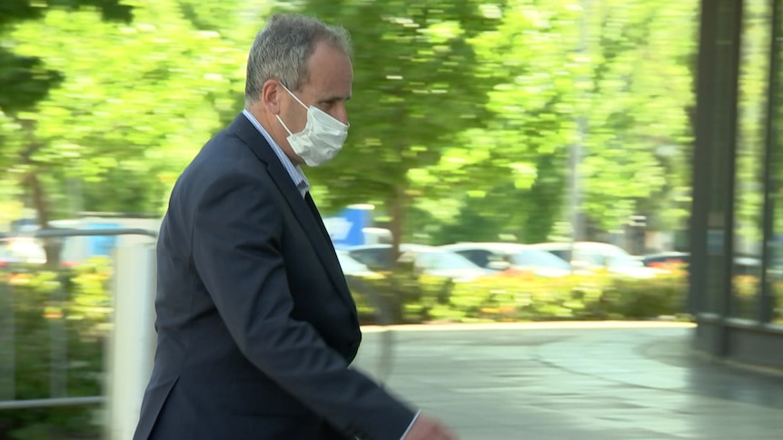 A man in a suit and wearing a mask, walking in front of a court building.