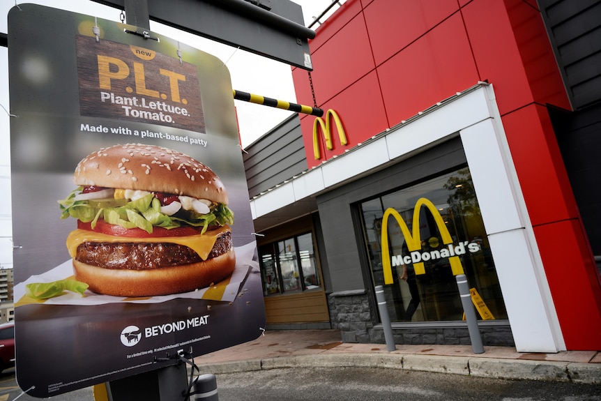 A sign with a picture of a delicious-looking burger is attached to a pole outside the entrance to a McDonald's restaurant.