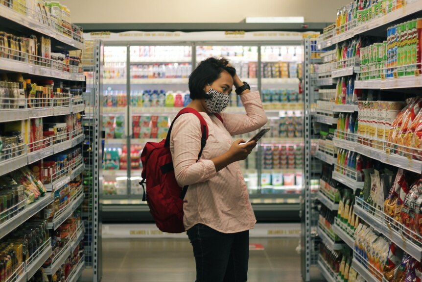 A woman wearing a mask and backpack looks at her phone and clutches her hair in a supermarket aisle.