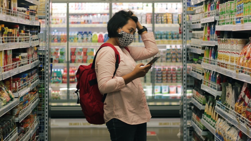 Woman wearing a mask and backpack looks at her phone and clutches her hair in a supermarket, in story about gluten free items.