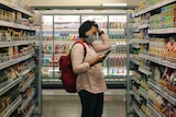 Woman wearing a mask and backpack looks at her phone and clutches her hair in a supermarket, in story about gluten free items.
