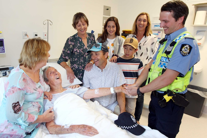 A police officer shakes hands with an elderly woman in a hospital bed, surrounded by family members.
