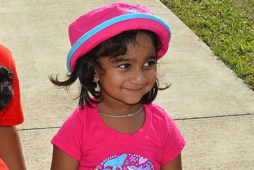 A tight mid-shot of a young Tamil girl wearing a pink shirt and pink and blue hat.