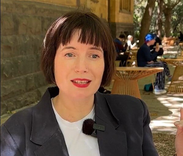 A white woman with a dark haired bob and red lipstick, in an outdoor space with people people sitting the background.
