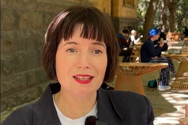 A white woman with a dark haired bob and red lipstick, in an outdoor space with people people sitting the background.