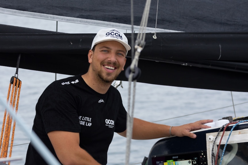 A smiling man wearing a white cap and black tshirt and shorts sits on a boat