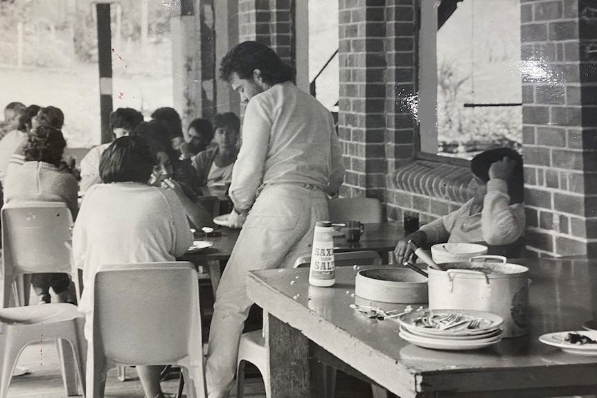 Lots of people gather at a table to eat food, a man dressed in 1970s flared tight jeans 