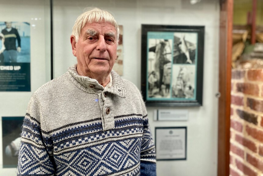 Elderly man standing in front of cabinet with historic photos wearing blue and grey knit jumper