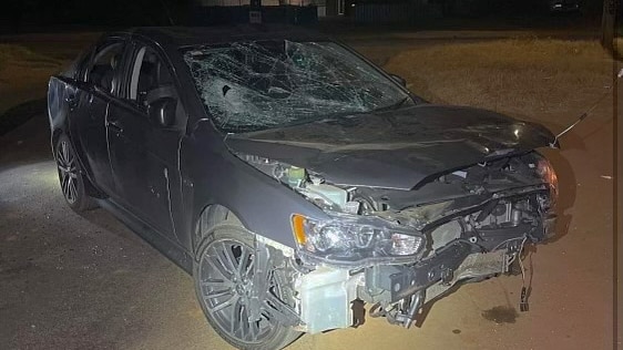 A grey sedan sits abandoned on the verge at night, its front end dented, windows smashed.