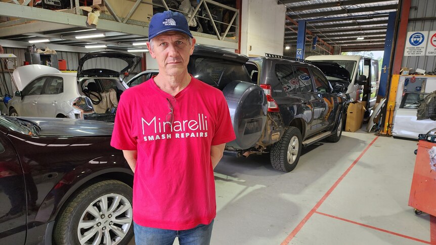A man in a red t-shirt and blue cap stands in a workshop full of cars.