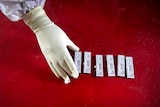 From above, you view a gloved hand and arm in full PPE taking completed nasal swab samples on a blood red table.