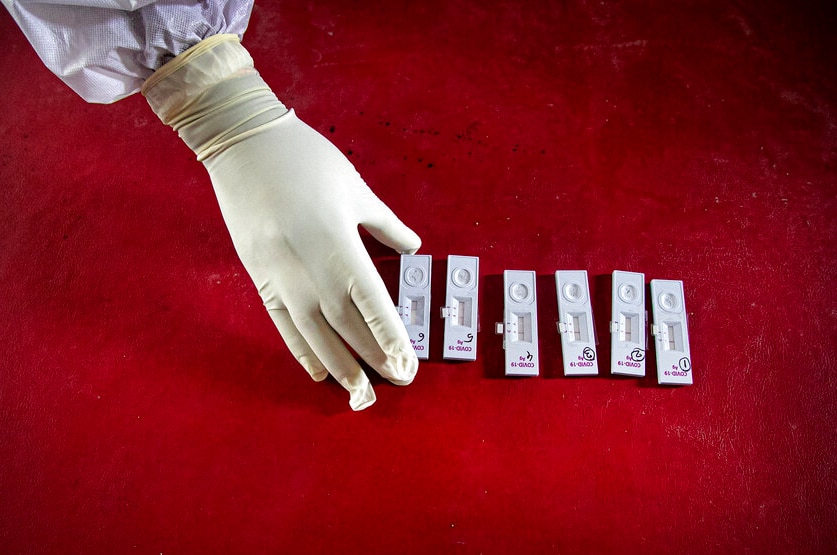 From above, you view a gloved hand and arm in full PPE taking completed nasal swab samples on a blood red table.