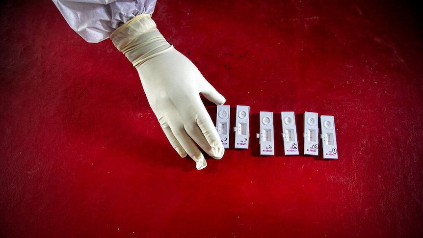 From above, you view a gloved hand and arm in full PPE taking completed nasal swab samples on a blood red table.