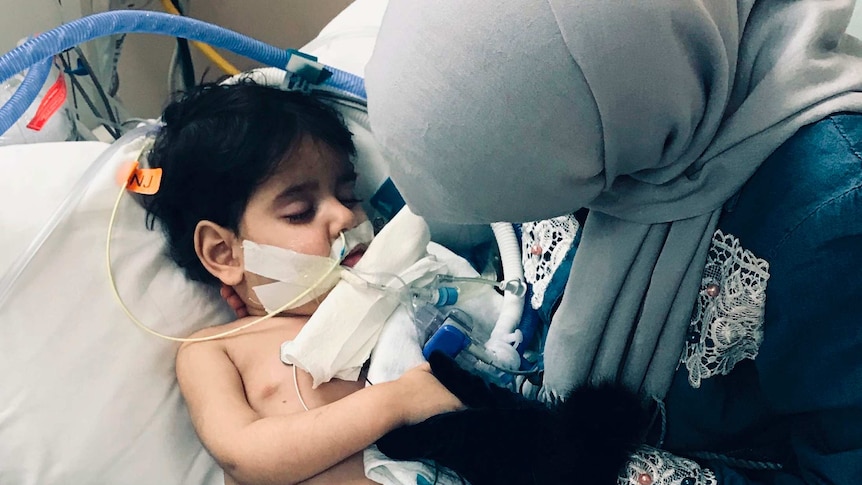 A woman in a headscarf holds her drying son on a hospital bed connected to breathing apparatus.