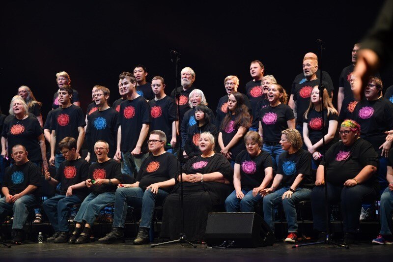choir of men and women in black tshirts with coloured circles on chest both seated and standing singing 