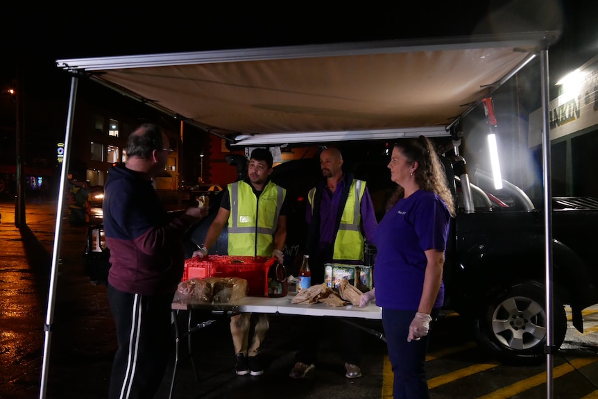 Four people stand at a food-covered table under an awning in the dark.