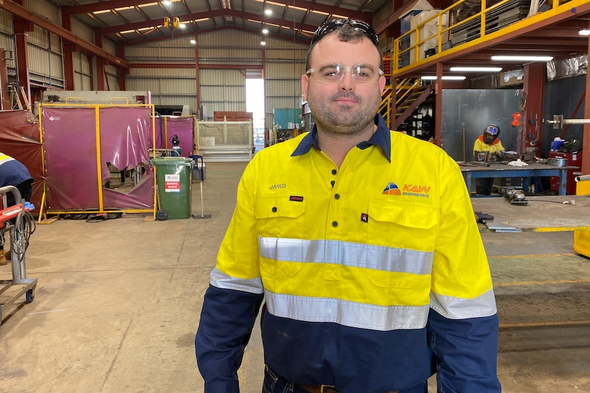 A man wearing a bight yellow high-vis short stands in the middle of a large warehouse, staring at the camera