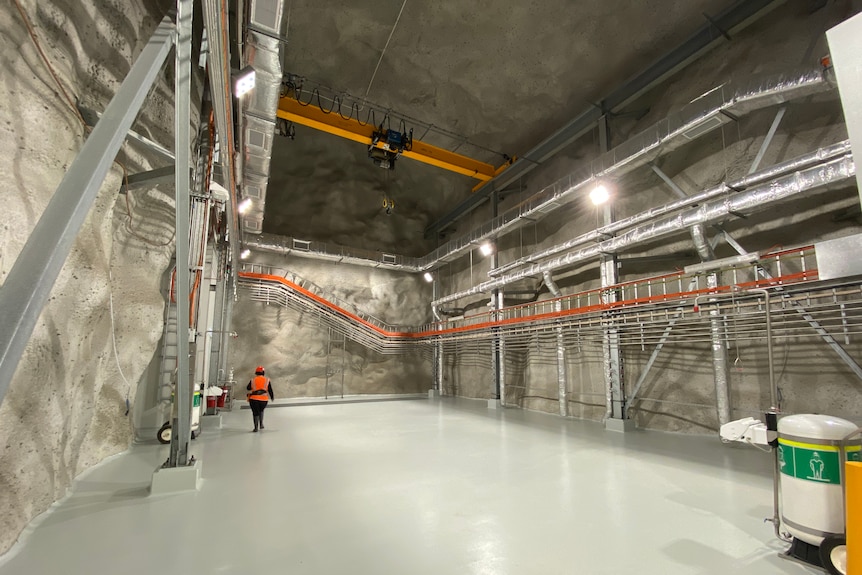 A large underground room where the walls are carved out of rock and pipes line the walls.