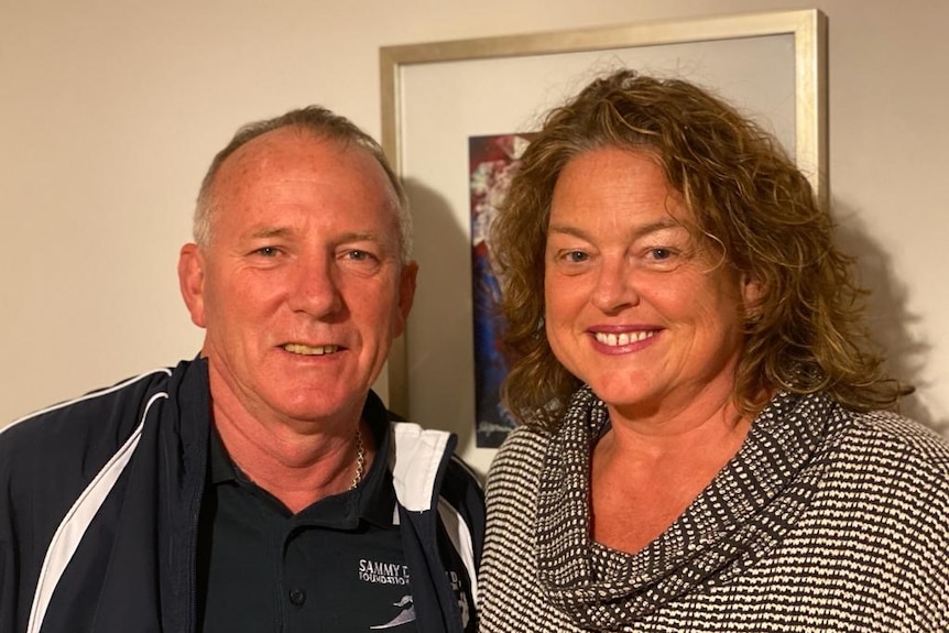 A middle aged man stands to the right of a middle aged woman, who has curly hair. They are inside, in front of a picture frame.