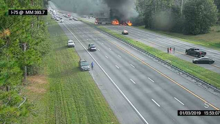 A highway showing cars stopped and people watching a firey crash on the side of the road