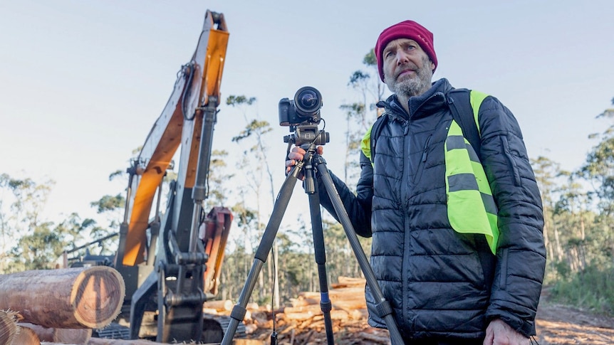 A man with photographic equipment stands next to logging machinery.