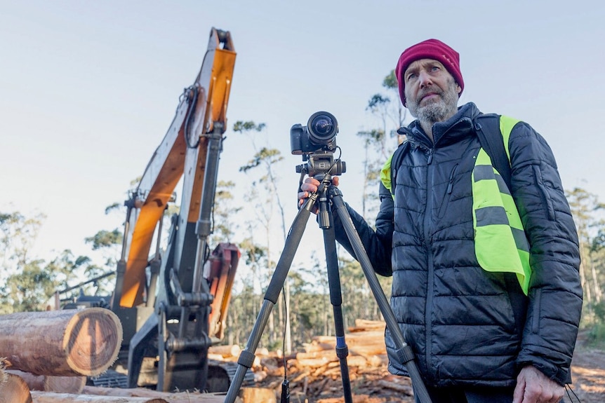 A man with photographic equipment stands next to logging machinery.