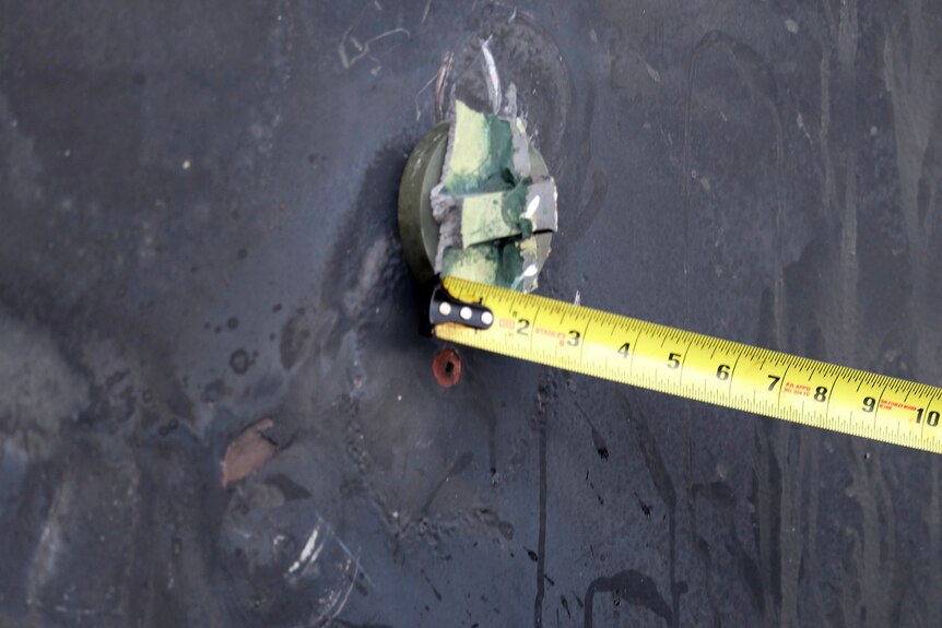 Yellow measuring tape is placed beside a circular piece of metal attached to what appears to be a ship hull.