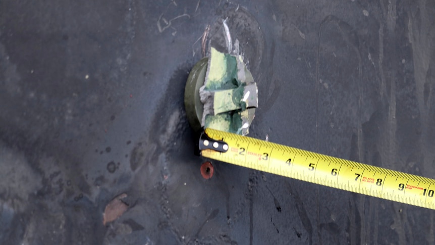 Yellow measuring tape is placed beside a circular piece of metal attached to what appears to be a ship hull.