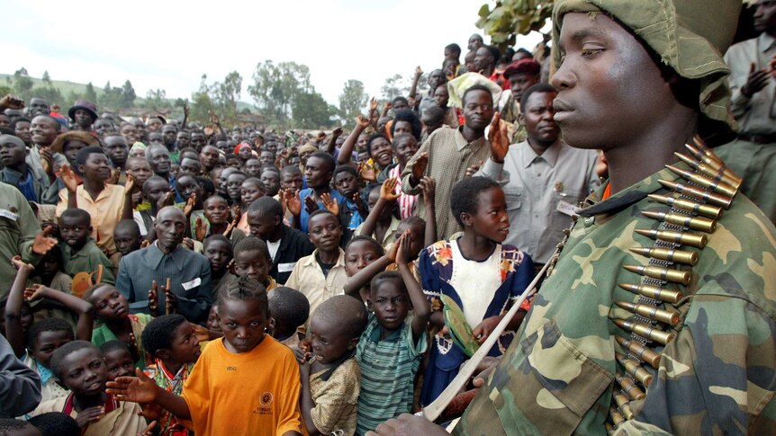 An armed soldier with an AK-47 and magazine stands in front of hoards of children posing for the camera.
