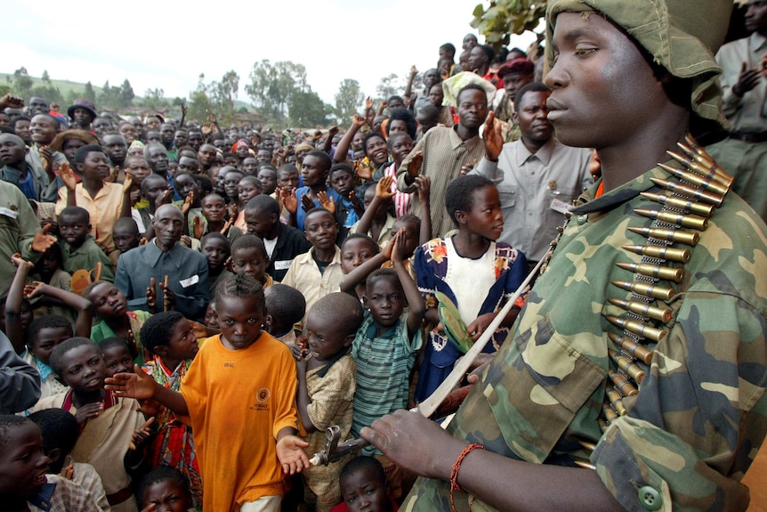 An armed soldier with an AK-47 and magazine stands in front of hoards of children posing for the camera.