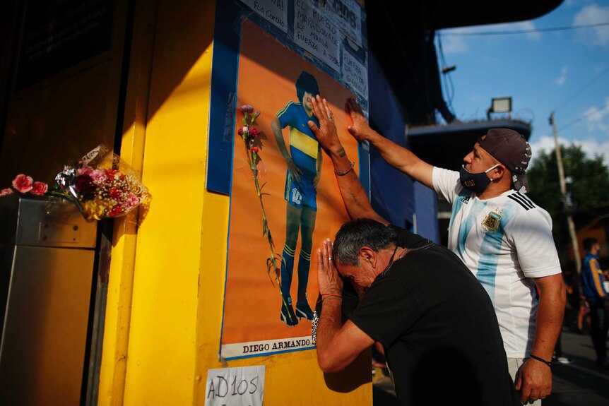 Two people touch a poster of Diego Maradona in his Boca Juniors kit. One is crying up against the wall
