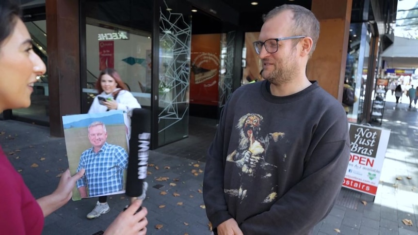 Man on the street looks at a photo being held by a reporter who is holding a microphone.