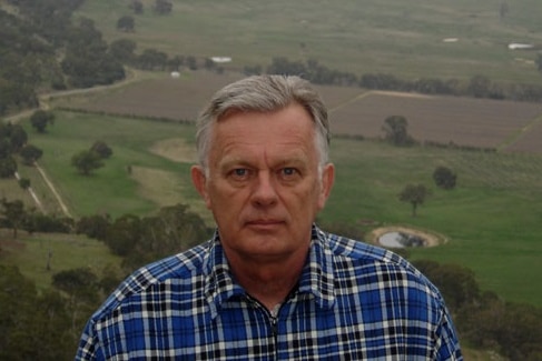 Broderick Smith, wearing a plaid shirt and with short grey hair, looking at the camera while standing outside.