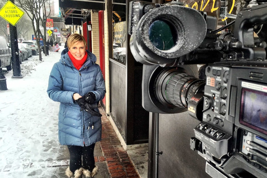 A blonde reporter stands in front of a camera as snow falls on a small town street in America