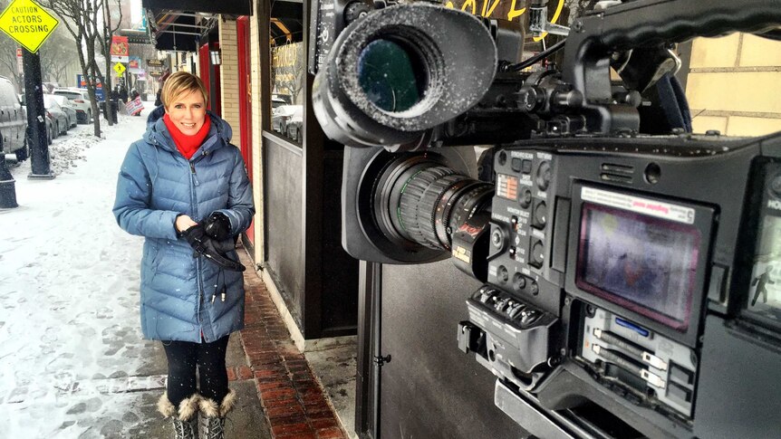 A blonde reporter stands in front of a camera as snow falls on a small town street in America