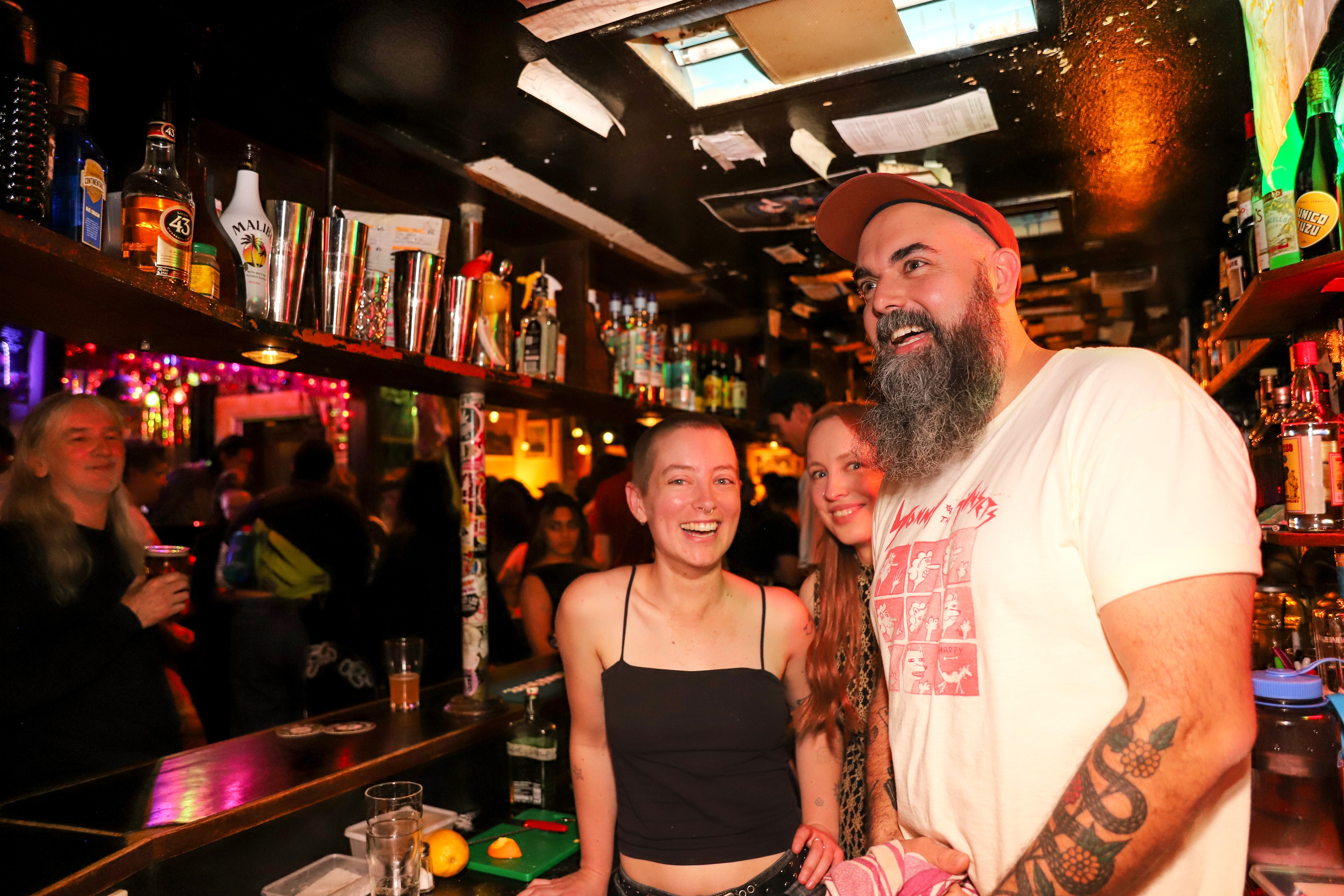 Três funcionários do bar sorriem atrás do bar em um lugar lotado à noite