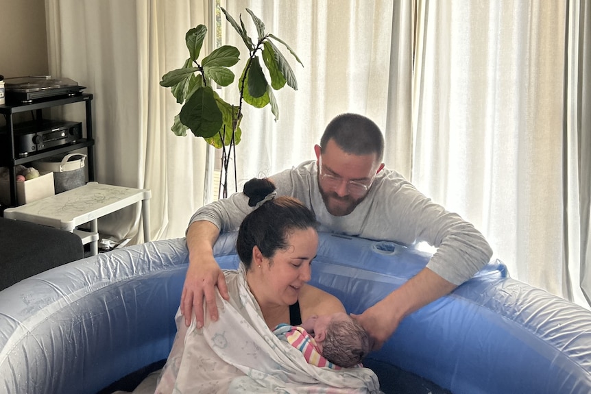 A woman in a pool inside a house holding her baby with a man embracing her