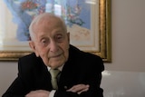 Close-up photo of Abram Goldberg in suit and tie, leaning folded arms across a table, smiling very slightly.