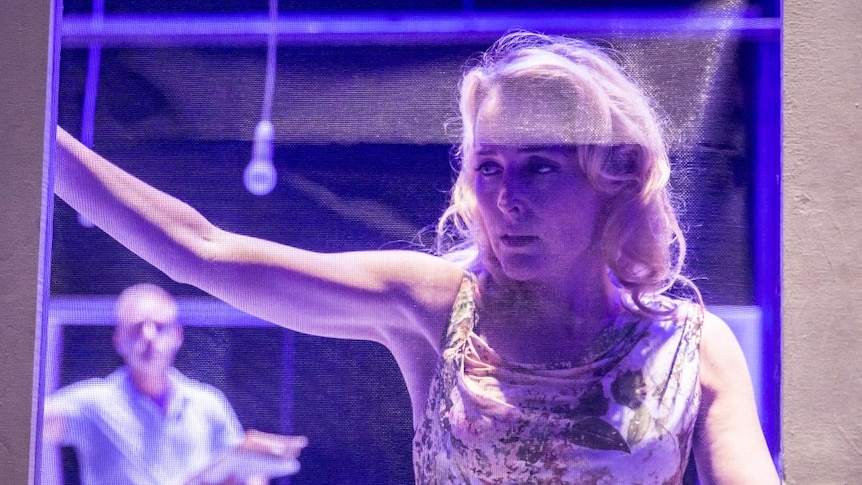 Woman with blonde hair seen on blue-lit stage, through screen mesh.