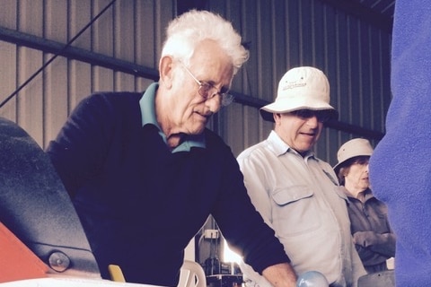 A bespectacled older man with a shock of white hair stands in a shed of some kind.