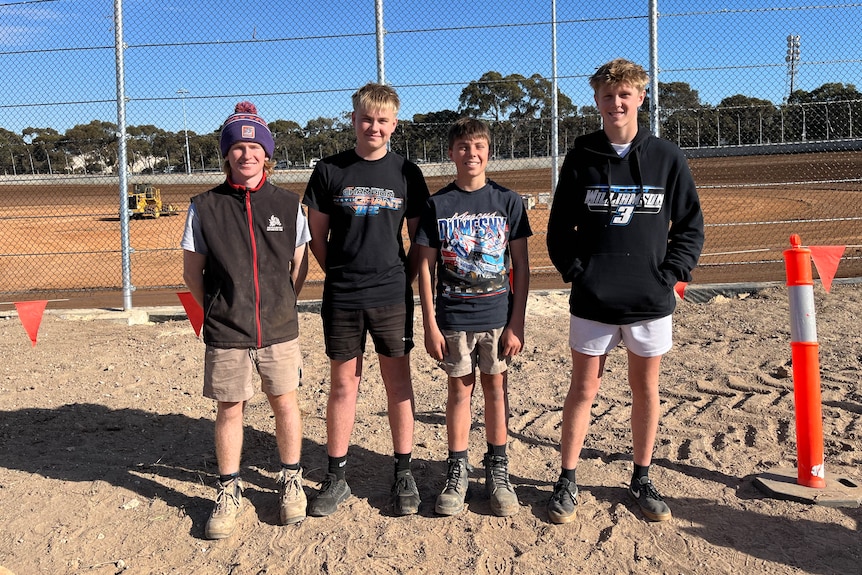 Four boys stand in front of a dirt racetrack