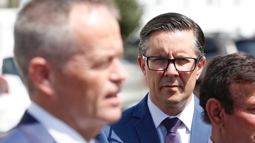 Butler wearing dark-rimmed glasses watches an out of focus Shorten speak to the media.